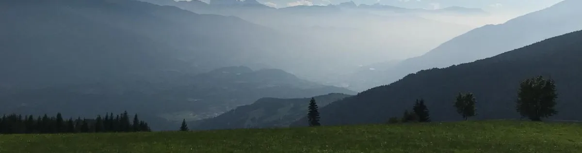 Bergwiese, dahinter ein Tal im Dunst und ferne Gipfel im Gegenlicht