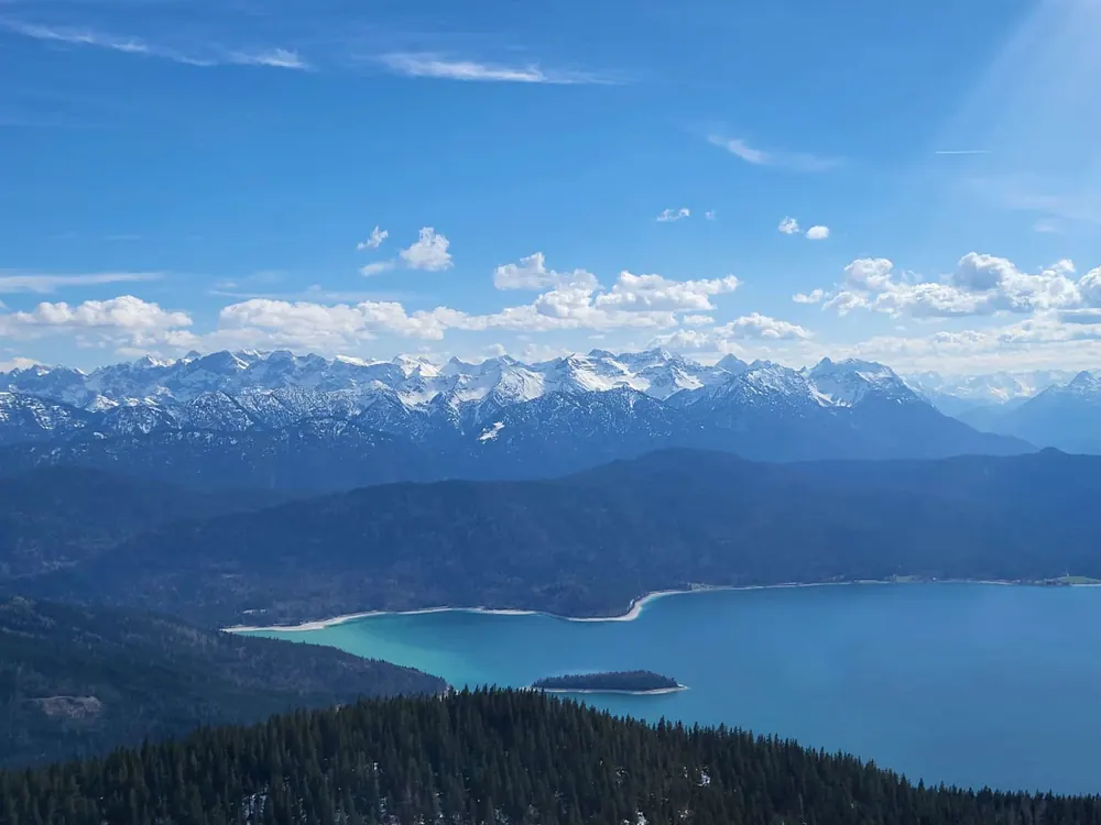 Weiter Blick über einen türkisfarbenen Bergsee auf eine schneebedeckte Alpenkette
